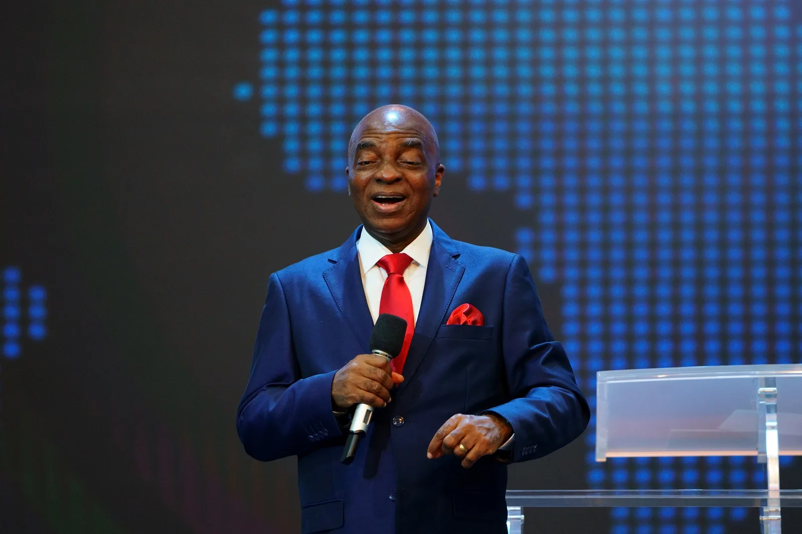 Bishop David Oyedepo, Founder and Presiding Bishop of Living Faith Church Worldwide, preaching in a blue suit with a microphone, a lit world map on the screen behind him
